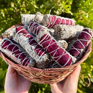 May include: A woven basket filled with sage bundles, some wrapped with dried rose petals and white string. The sage bundles are a light gray color, and the rose petals are a deep red. The basket is made of light brown woven material.