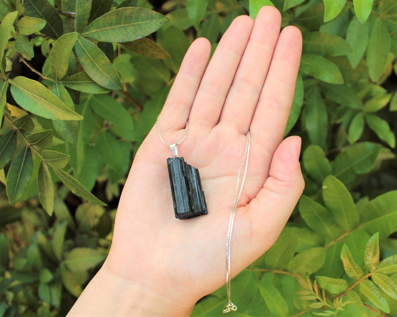 May include: A black tourmaline pendant necklace with a silver chain. The tourmaline is a dark, opaque stone with a rough, textured surface. The necklace is held in a hand against a backdrop of green leaves.