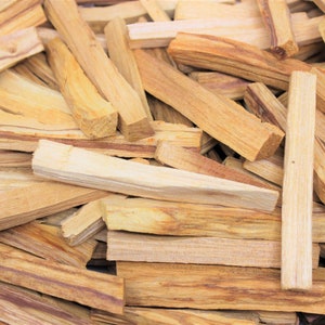 May include: A collection of light brown Palo Santo wood sticks. The sticks are rectangular, with varying shades of tan and brown. Some sticks have visible grain patterns and knots. The wood appears to be freshly cut.