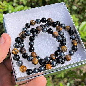 Pode incluir: Duas pulseiras de contas, uma com contas de olho de tigre e a outra com contas pretas e de hematita, s&atilde;o exibidas em uma caixa de presente branca.