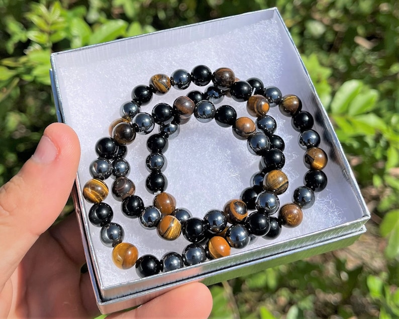 Pode incluir: Duas pulseiras de contas, uma com contas de olho de tigre e a outra com contas pretas e de hematita, s&atilde;o exibidas em uma caixa de presente branca.