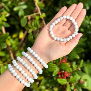 May include: Four white beaded bracelets stacked on a person's wrist. The bracelets are made of small, round beads with a white and gray marbled pattern.