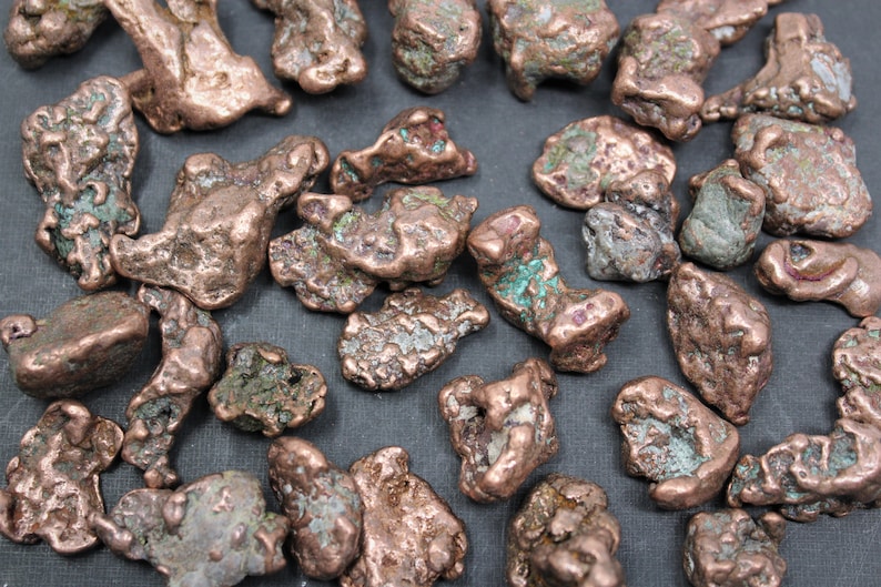 May include: A collection of raw copper nuggets featuring irregular, textured shapes with a metallic reddish-brown finish and patches of green oxidation. These natural mineral specimens display varied sizes and jagged, organic forms throughout.