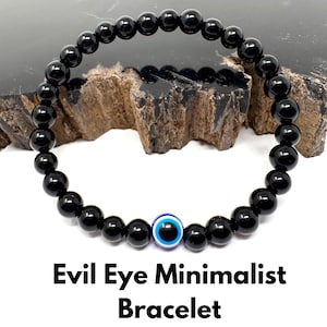 May include: Black beaded bracelet with an Evil Eye charm. The bracelet features small, round, polished black beads strung together. The Evil Eye charm is blue and white. The text "Evil Eye Minimalist Bracelet" is at the bottom.
