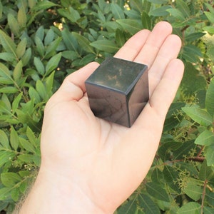 May include: A dark green, almost black, cube-shaped crystal. The crystal is smooth and polished, with a slight sheen. It is being held in a person's hand.