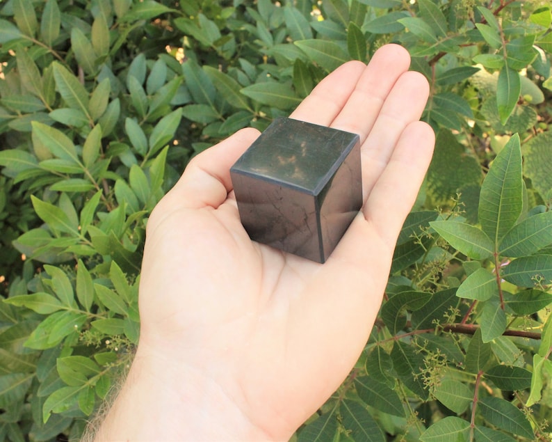 May include: A dark green, almost black, cube-shaped crystal. The crystal is smooth and polished, with a slight sheen. It is being held in a person's hand.