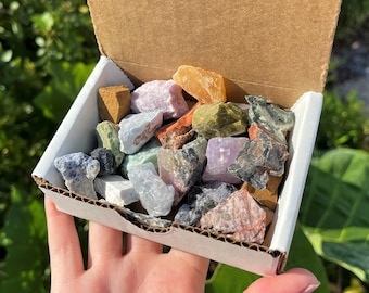 May include: A cardboard box filled with a variety of colorful tumbled stones, including green, blue, purple, red, and orange. The stones are arranged in a random pattern and are all different sizes.