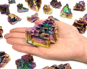 May include: A hand holding a large, iridescent bismuth crystal with a geometric, stepped structure. The crystal displays a vibrant array of colours, including blues, greens, yellows, and purples. Other smaller crystals are scattered around on a white surface.