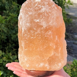 May include: A large, natural Himalayan salt lamp with a warm orange hue. The lamp is sitting on a wooden base and is being held in a person's hand.