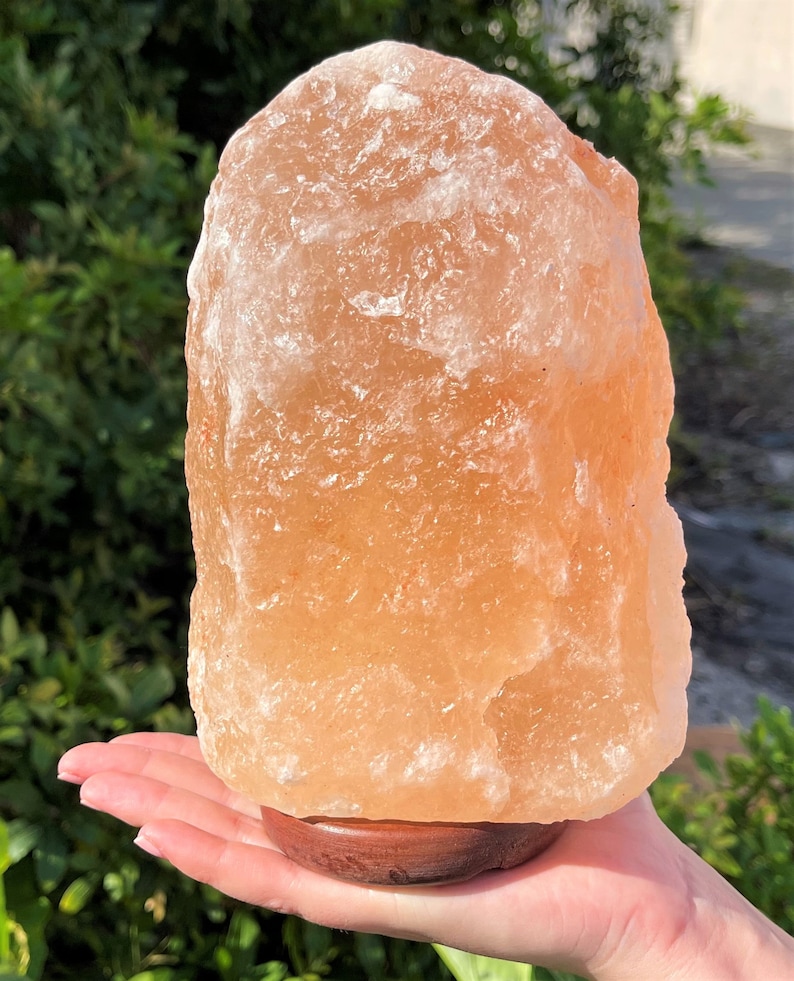 May include: A large, natural Himalayan salt lamp with a warm orange hue. The lamp is sitting on a wooden base and is being held in a person's hand.