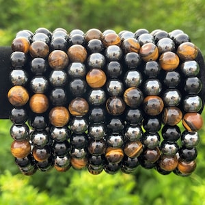 Pode incluir: Uma pilha de pulseiras feitas com contas de olho de tigre e hematita. As pulseiras s&atilde;o de cor marrom, dourado, preto e prateado.