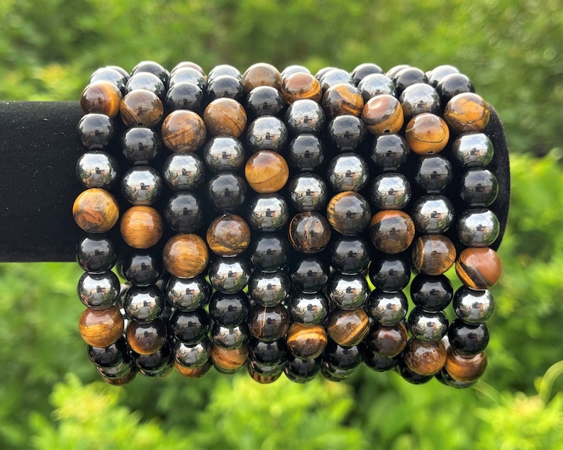 Pode incluir: Uma pilha de pulseiras feitas com contas de olho de tigre e hematita. As pulseiras s&atilde;o de cor marrom, dourado, preto e prateado.