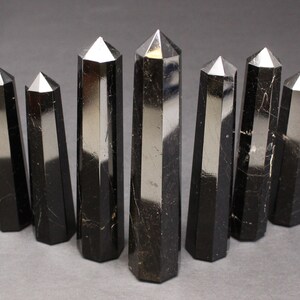 May include: Eight black obsidian crystal points, also known as black glass, are arranged in a row on a gray surface. The points are all roughly the same size and shape, and they have a smooth, polished finish.