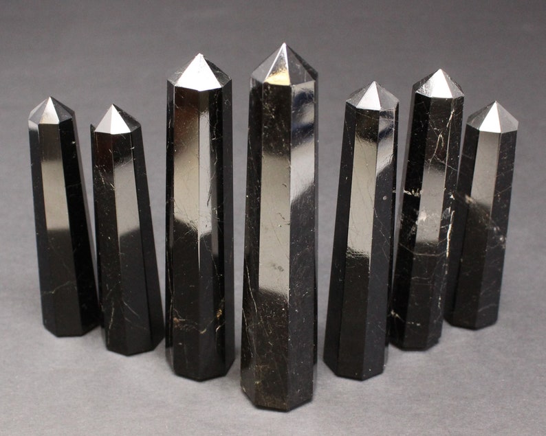 May include: Eight black obsidian crystal points, also known as black glass, are arranged in a row on a gray surface. The points are all roughly the same size and shape, and they have a smooth, polished finish.