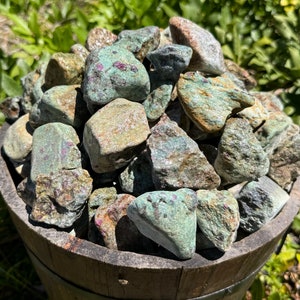 May include: A collection of green and pink tumbled stones in a wooden bucket. The stones are smooth and have a variety of shapes and sizes.