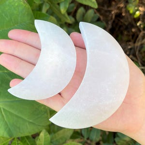 May include: Two crescent moon-shaped objects carved from translucent white selenite crystal. The two pieces feature a smooth, polished surface with a subtle crystalline texture and a bright, opaque white colour throughout the curved forms.