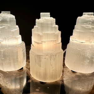 May include: Three white selenite tower lamps with a layered design are illuminated against a dark background. The lamps are cylindrical, with a textured, crystalline appearance. The lamps are lit from within, creating a soft glow.