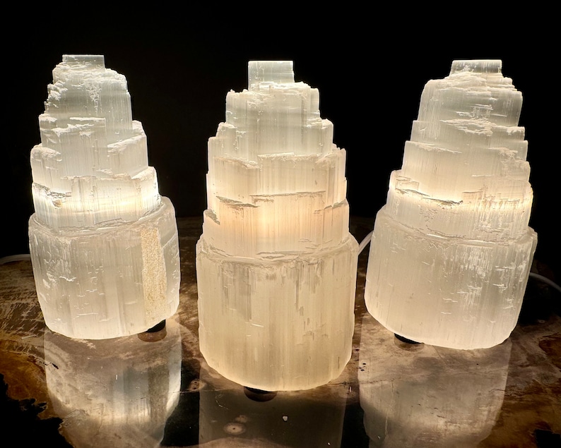 May include: Three white selenite tower lamps with a layered design are illuminated against a dark background. The lamps are cylindrical, with a textured, crystalline appearance. The lamps are lit from within, creating a soft glow.