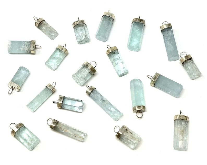 May include: A collection of aquamarine crystal pendants with silver-colored caps and loops. The pendants are various shapes and sizes, displaying a pale blue-green color. The background is white, highlighting the pendants.