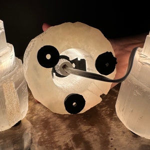 May include: A selenite lamp with a circular base and a cylindrical top. The lamp is made of a translucent, off-white material and has a black power cord. The lamp is lit from within, creating a soft glow.