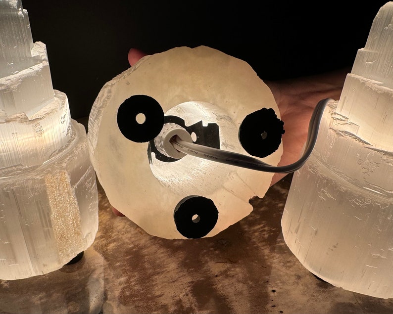 May include: A selenite lamp with a circular base and a cylindrical top. The lamp is made of a translucent, off-white material and has a black power cord. The lamp is lit from within, creating a soft glow.