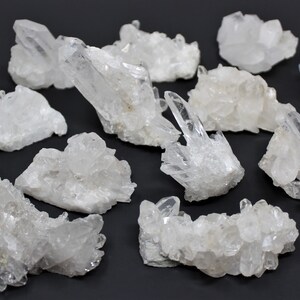 May include: A collection of 15 clear quartz crystal clusters. The clusters are various sizes and shapes, with some having a more pointed shape and others being more rounded. The crystals are all clear and have a natural, unpolished look.