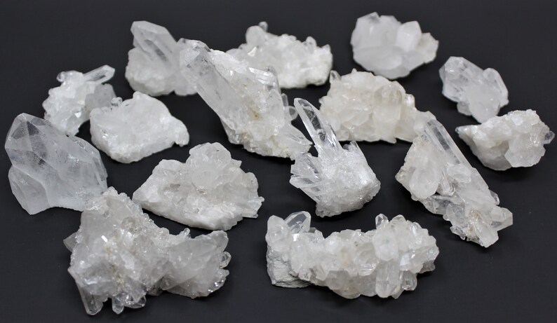 May include: A collection of 15 clear quartz crystal clusters. The clusters are various sizes and shapes, with some having a more pointed shape and others being more rounded. The crystals are all clear and have a natural, unpolished look.