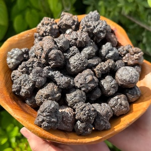 May include: A wooden bowl filled with a collection of dark, textured hematite stones. The stones have a rough, irregular surface and a metallic sheen. The bowl is a warm, natural wood color, and the background is blurred green foliage.