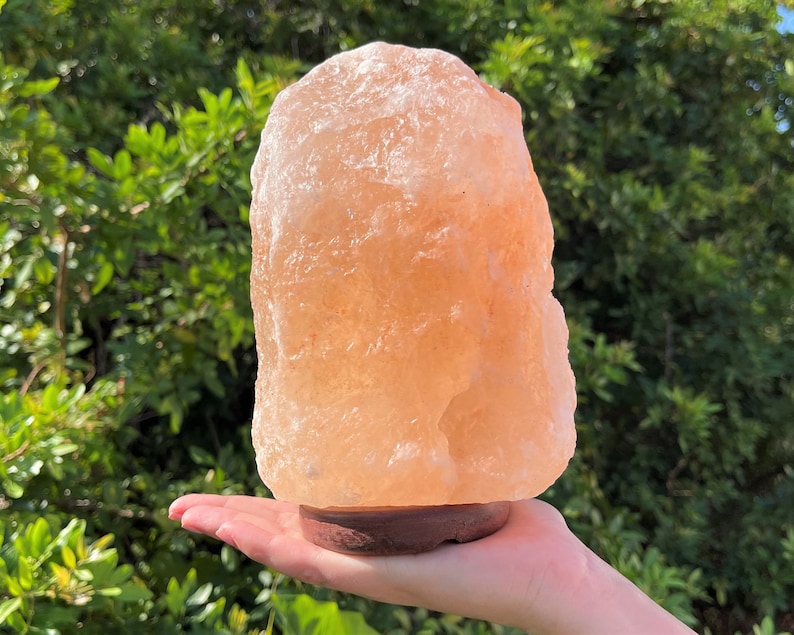 May include: A large, natural Himalayan salt lamp with a warm, orange-pink hue. The lamp is sitting on a wooden base and is lit, casting a soft glow.