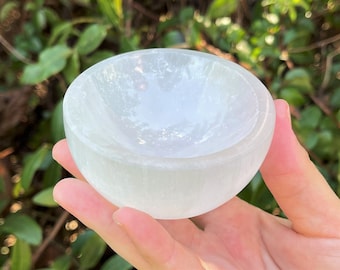 May include: A white selenite bowl with a smooth, rounded shape. The bowl is held in a hand, showcasing its translucent quality. The bowl is designed for crystals, meditation, or home decor.