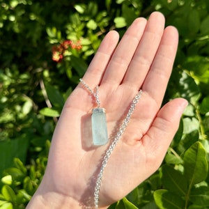 May include: A silver necklace featuring a rectangular, pale blue aquamarine pendant. The pendant is suspended from a delicate silver chain. The background is a blurred view of green foliage.