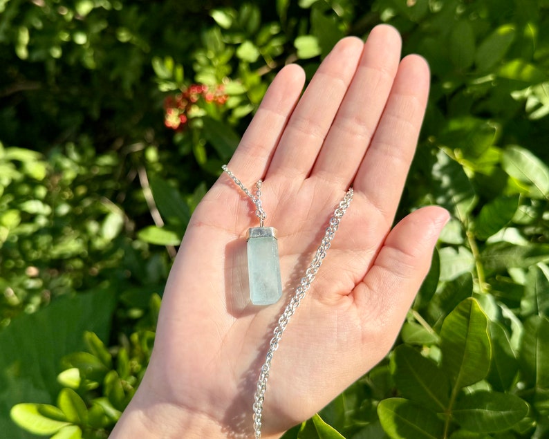 May include: A silver necklace featuring a rectangular, pale blue aquamarine pendant. The pendant is suspended from a delicate silver chain. The background is a blurred view of green foliage.