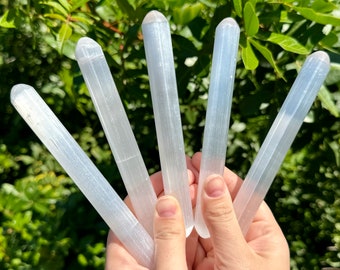 May include: Five white selenite wands with a smooth, polished finish. The wands are held in a hand, showcasing their length and shape.