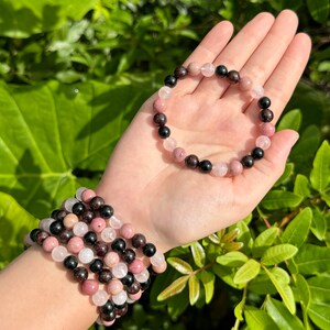 May include: A bracelet made of pink, black, and red gemstone beads. The bracelet is shown on a person's wrist and in the palm of a hand.