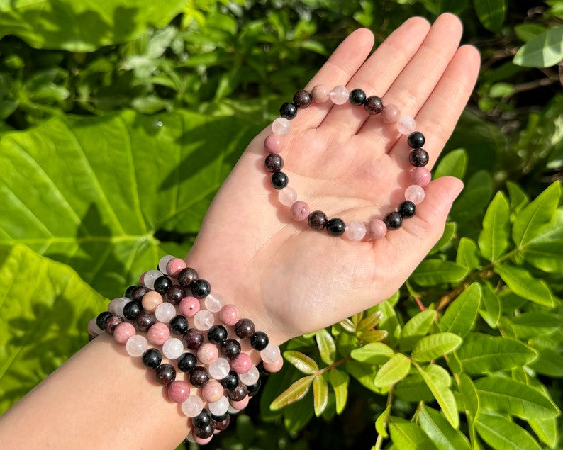 May include: A bracelet made of pink, black, and red gemstone beads. The bracelet is shown on a person's wrist and in the palm of a hand.