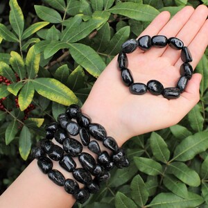 May include: A set of two bracelets made of polished, irregular black stone beads. One bracelet is worn around the wrist, consisting of multiple stacked strands of these smooth, dark, rounded stones, while the other is held in the palm of the hand.