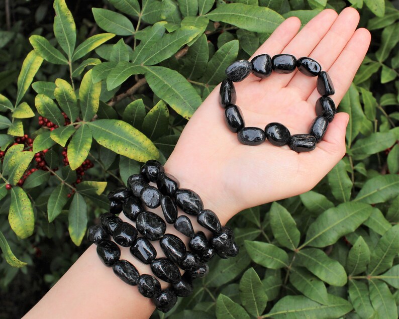 May include: A set of two bracelets made of polished, irregular black stone beads. One bracelet is worn around the wrist, consisting of multiple stacked strands of these smooth, dark, rounded stones, while the other is held in the palm of the hand.