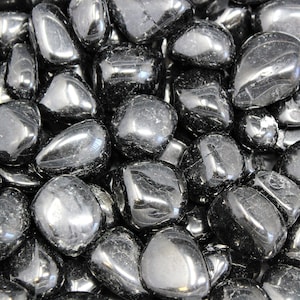 May include: A close-up view of a pile of black obsidian tumbled stones. The stones are smooth and polished, with a glossy finish.