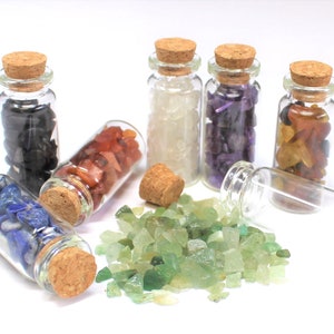 May include: Seven small glass bottles with cork stoppers, each filled with different colored crystals. The bottles are arranged in a semi-circle on a white surface. The crystals are: black, white, purple, orange, red, blue, and green.