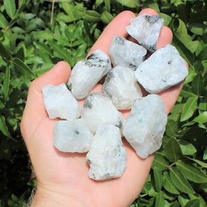 May include: A collection of white rainbow moonstone rough crystals with black and green inclusions. The crystals are various sizes and shapes.