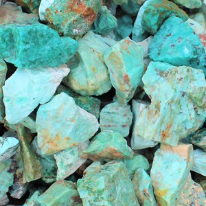 K&ouml;nnte beinhalten: Eine Ansammlung von grob geschnittenen Amazonitsteinen in T&uuml;rkis-, Gr&uuml;n- und Blaut&ouml;nen. Die Steine haben ein nat&uuml;rliches, strukturiertes Aussehen mit Farbvariationen und einigen braunen und wei&szlig;en Bereichen. Das Bild ist eine Nahaufnahme.