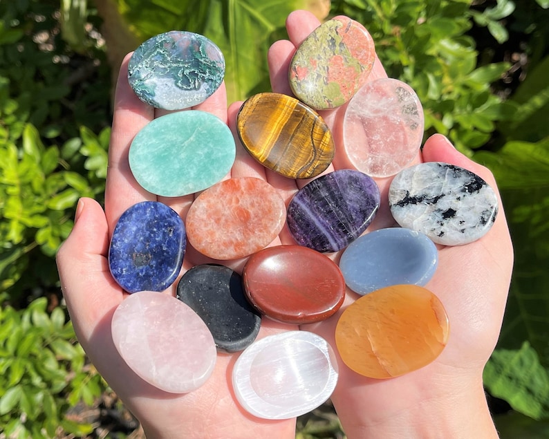 May include: A collection of 14 smooth, polished, oval-shaped stones in various colours, including green, blue, red, orange, purple, and white. The stones are arranged in a circle on a person's hands.