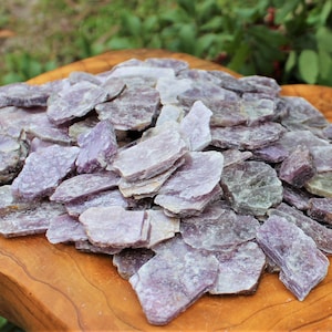 May include: A large pile of purple and gray mica flakes, also known as muscovite, on a wooden surface. The flakes are translucent and have a shimmering effect.