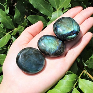 May include: Three smooth, polished labradorite stones in shades of blue and green with iridescent flashes. The stones are arranged in a row on a hand.