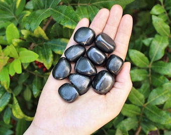 May include: A collection of black tumbled stones, likely hematite, in a hand. The stones are smooth and polished, with a slightly metallic sheen.