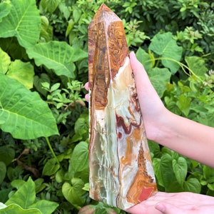 May include: A tall, polished stone obelisk featuring a complex pattern of swirling bands in shades of cream, translucent pale green, deep brown, and rust red. The crystal tower has a pointed apex and a smooth, multi-coloured surface texture.