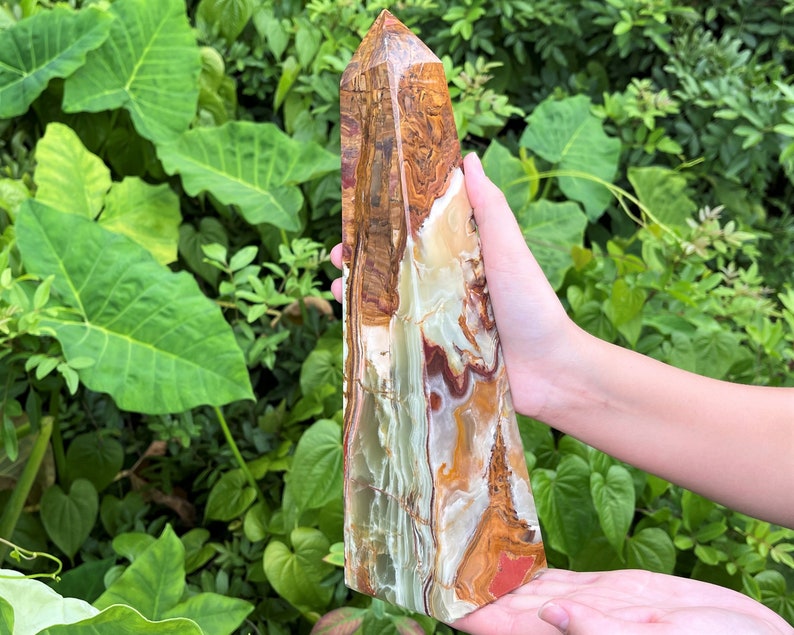 May include: A tall, polished stone obelisk featuring a complex pattern of swirling bands in shades of cream, translucent pale green, deep brown, and rust red. The crystal tower has a pointed apex and a smooth, multi-coloured surface texture.