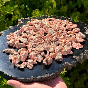May include: A collection of raw, metallic copper nuggets with a shiny, reddish-brown finish piled on top of a flat, circular, polished black stone slab with a rough, natural outer edge, displaying a high-contrast texture and color palette.