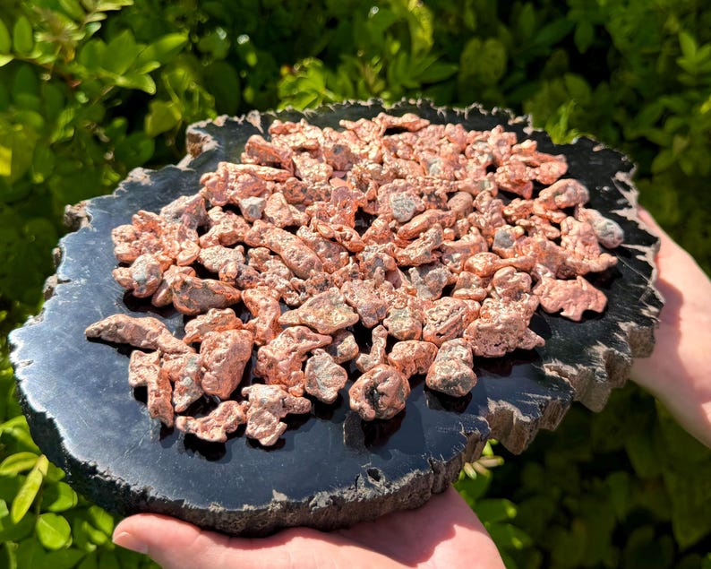 May include: A collection of raw, metallic copper nuggets with a shiny, reddish-brown finish piled on top of a flat, circular, polished black stone slab with a rough, natural outer edge, displaying a high-contrast texture and color palette.