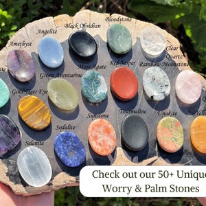 Op de afbeelding: Een assortiment van 16 worry- of palmstenen in verschillende kleuren en texturen, waaronder amethist, groene aventurijn, tijgeroog, sodaliet, rozenkwarts en helder kwarts. De stenen zijn in een cirkel op een houten oppervlak gerangschikt. De tekst "Ontdek onze 50+ unieke worry- en palmstenen" is zichtbaar onderaan de afbeelding.